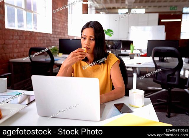 Caucasian businesswoman sitting at desk with coffee using laptop and biscuit