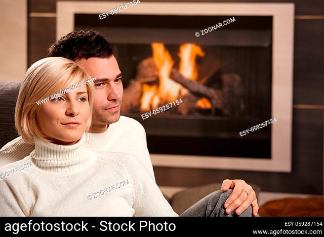 Young couple hugging on sofa in front of fireplace at home, looking away, smiling
