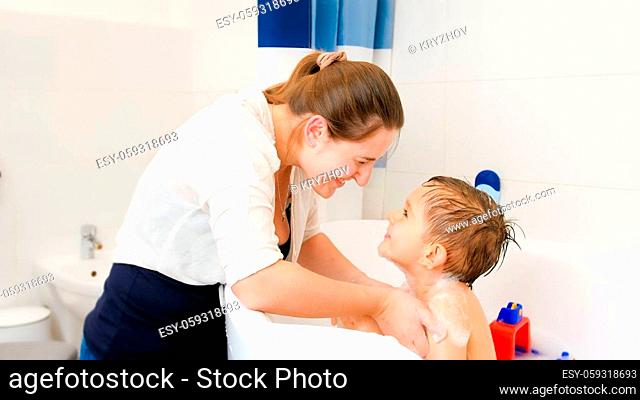 Smiling young caring mother washing her little boy sitting in bath with soap. Concept of child hygiene and health care at home