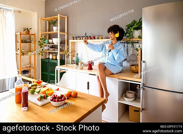 Smiling woman with coffee cup taking selfie through smart phone sitting on kitchen counter at home
