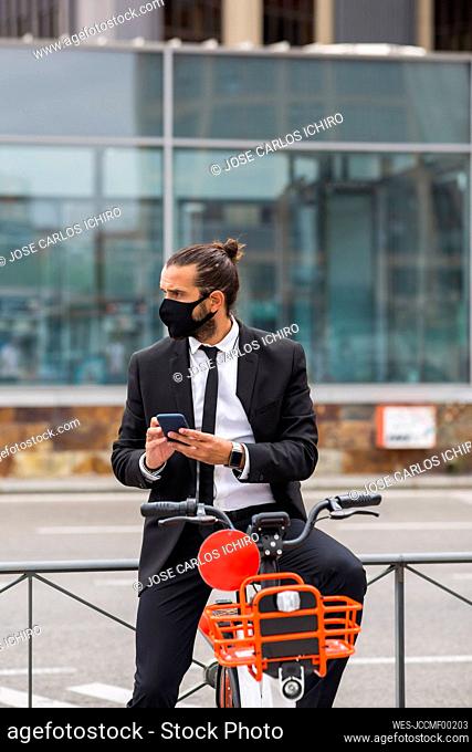 Portrait of businessman wearing protective face mask sitting on bicycle with smart phone in hands