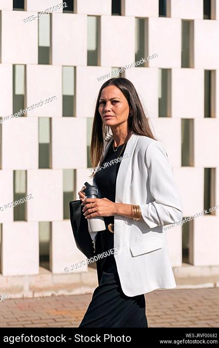Businesswoman with insulated drink container walking by wall