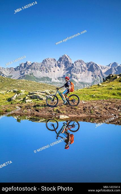 man, 22 years old, cyclist with e-bike near an alpine lake, cyclist reflected in the water of a mountain lake, mountains in the background