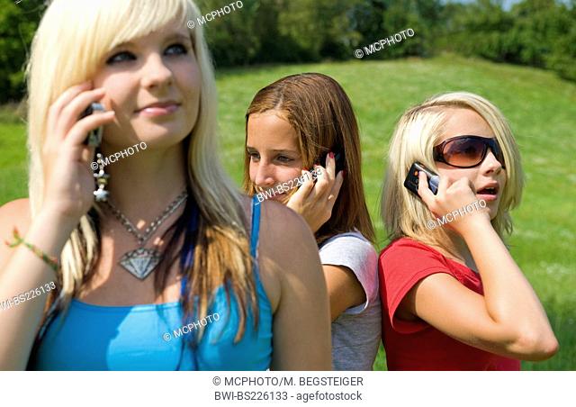 three young girls standing in a meadow phoning with a mobile with a smile