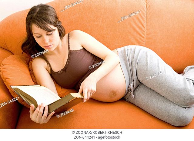Schwangere brünette Frau liegt auf einem orangen Sofa und liest ein Buch