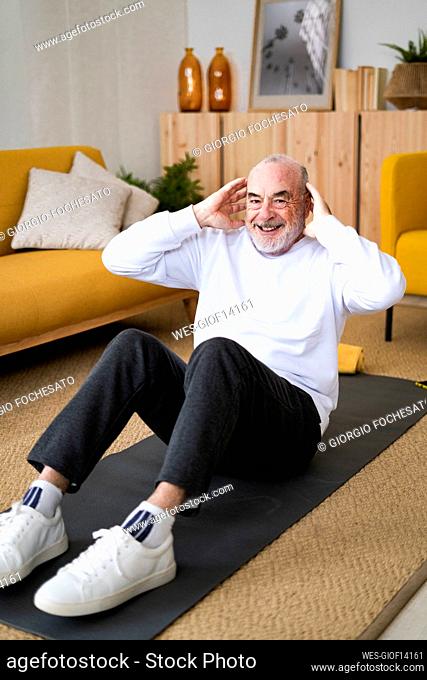 Smiling senior man with hands behind head exercising on mat at home