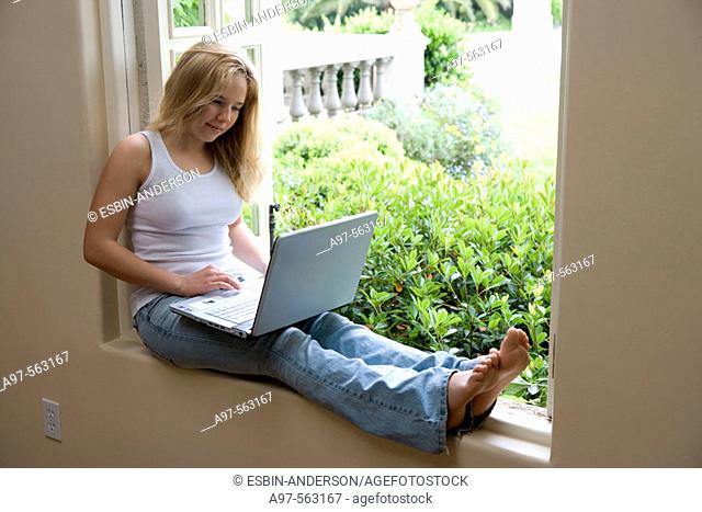 Blonde barefoot teen girl sits by an open window using her laptop computer