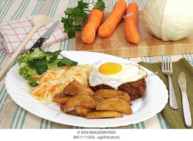 Farmers meat ball, hamburger steak, fried egg, baked potatoe wedges, coleslaw, carrots, white cabbage and flat leaf parsley on wooden board