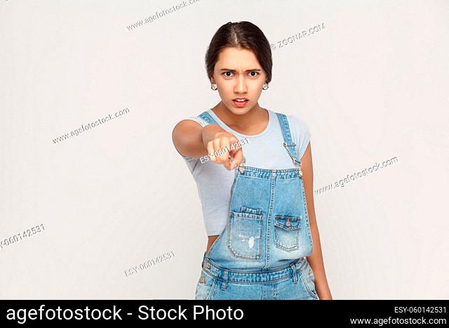 Angry latin woman with denim overalls looking at camera and pointing finger. Studio shot isolated on gray background