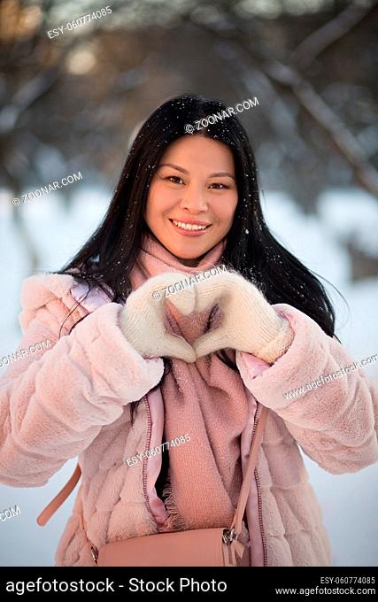 Beautiful Asian shows heart gesture, Winter Park on background. Smiling woman with flowing hair, dressed in a pink jacket and a mitten show love sign for...