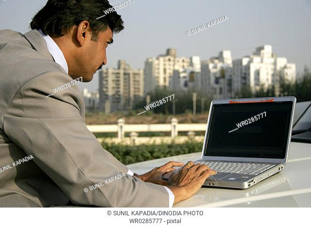 Indian businessman in gray coat and black pant using laptop on bonnet of car against some corporate buildings of Pune City , Maharashtra , India MR686B
