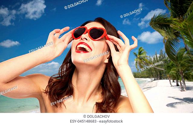 valentines day, beauty and people concept - happy smiling young woman with red lipstick and heart shaped sunglasses over exotic tropical beach with palm trees...