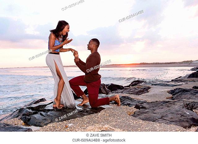Mid adult man kneeling on rocks beside sea, proposing to young woman