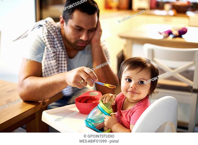 Portrait of baby girl sitting in high chair at home with annoyed father in background