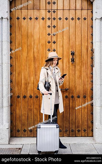Mid adult woman with wheeled luggage using smart phone against wood door