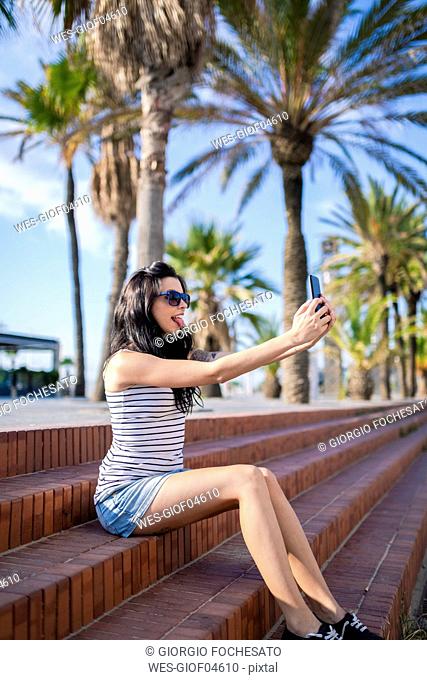 Spain, Barcelona, young woman sitting on steps at sunlight taking selfie with smartphone