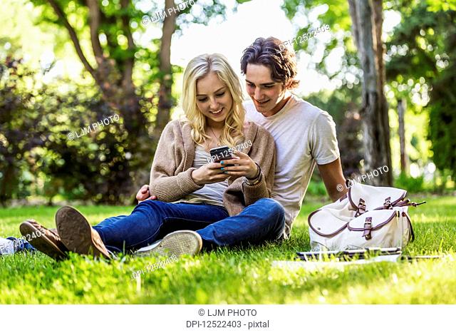 A young couple sits studying on the grass on the university campus with a textbook and tablet while using a smart phone; Edmonton, Alberta, Canada