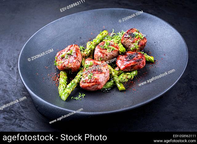 Traditional barbecue Iberian pork filet medaillons with green asparagus tips offered as close-up on a modern design cast iron black plate