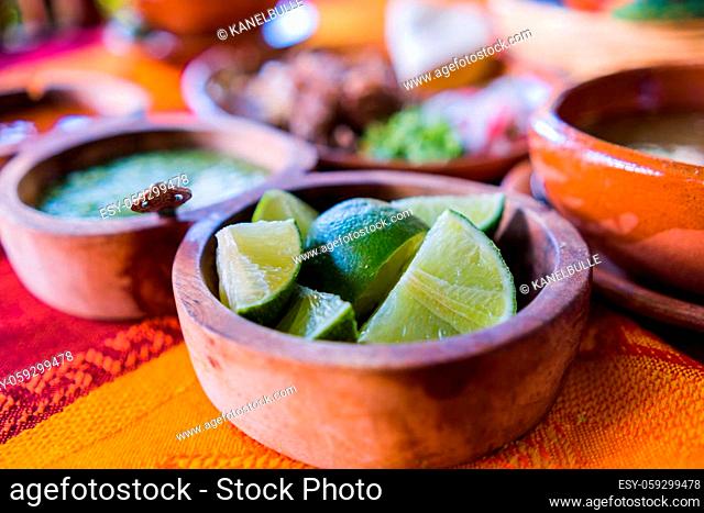 Close-up of clay bowls of lime slices, green sauce, and lamb broth with blurry chopped meat as background. Condiments and sliced vegetables above orange and red...