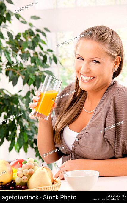 Young woman sitting at table with cereal and fruits, drinking orange juice, smiling