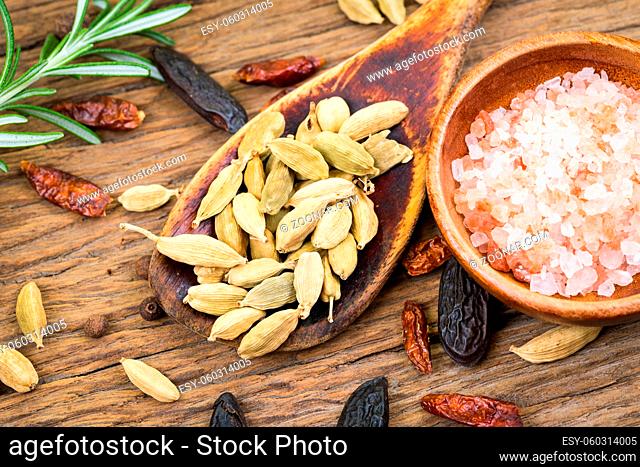 Closeup of whole cardamom on a wooden spoon and pink crystal salt in a small wooden bowl on rustic wood background and a sprig of rosemary