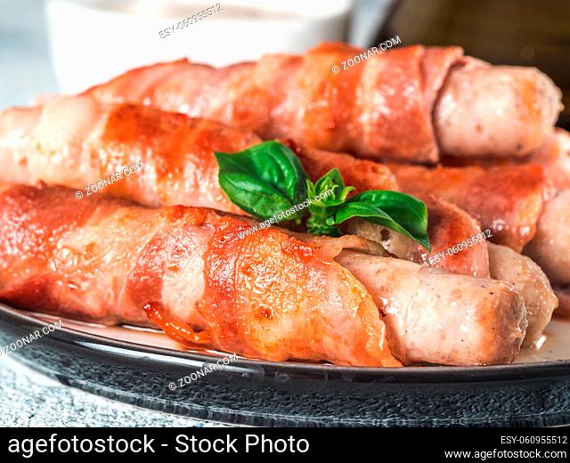 Ready-to-eat pigs sausages wrapped in bacon on plate. Fried savory sausages wrapped in bacon served fresh green basil leaves with sauce on background