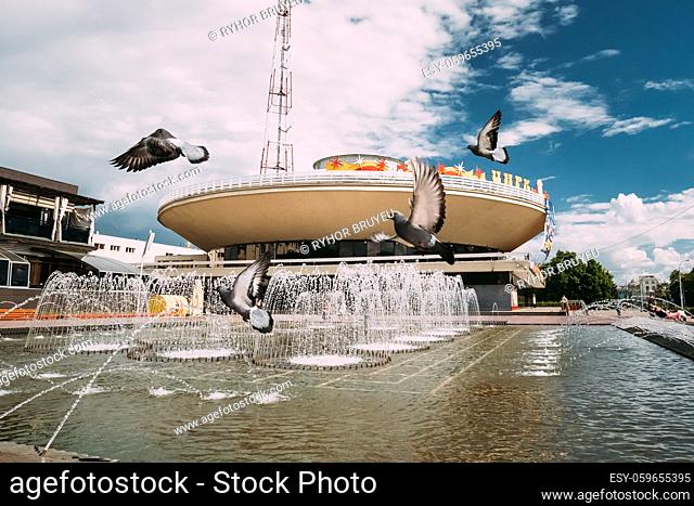 Gomel, Belarus. Pigeons Doves Are Flying Over A Fountain Near Gomel State Circus In A Summer Sunny Day. Famous Local Landmark Under Sunny Blue Sky In Summer Day
