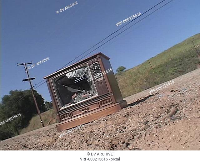 An abandoned vintage television set sits broken, with a shattered screen, on the side of a road