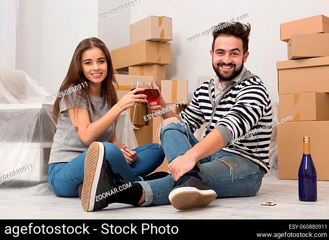 Moving in, time for joy, young couple celebrating relocation. Man and woman sitting on the floor in their new apartment, drinking wine and smiling