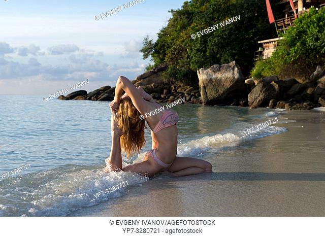 Yoga in the waves of Andaman sea - girl in pigeon pose practicing yoga at sunrise. Koh Lipe, Thailand