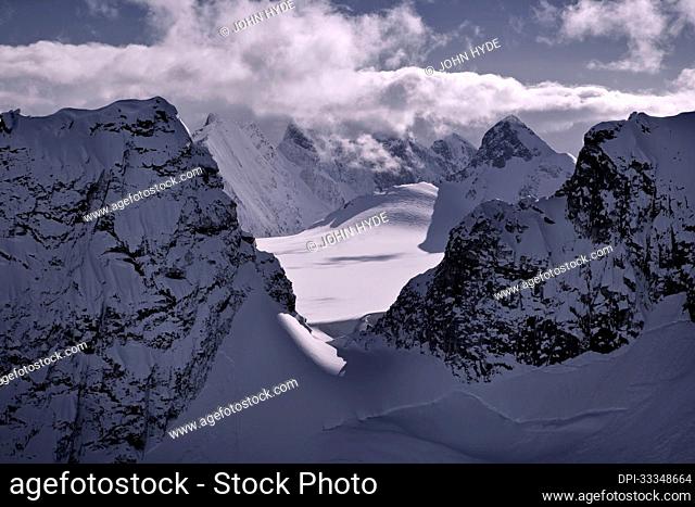 Snow covered jagged mountain peaks with a purple-grey hue surrounding the sunlit glaciers under a cloud bank on the Juneau Ice Field in the Tongass National...