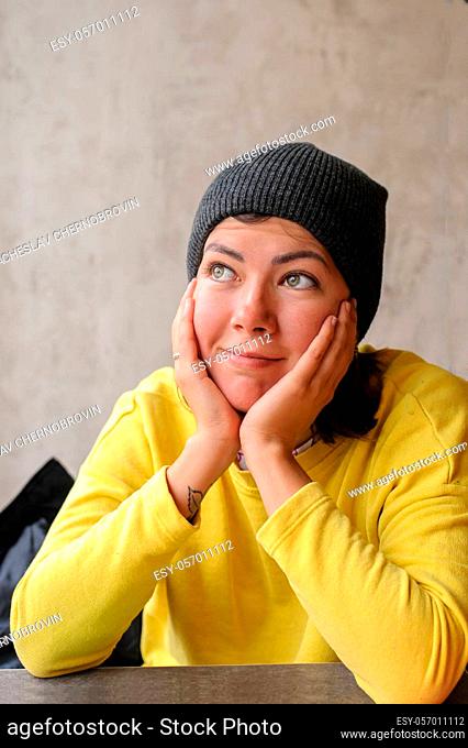 cute white young woman in yellow sweatshirt and knit cap smiling, resting her head on both her hands
