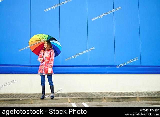Woman with multi colored umbrella standing on footpath by blue wall