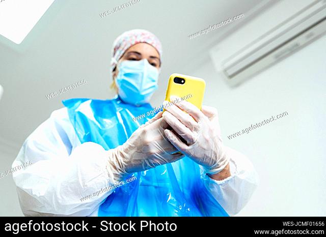 Dental doctor wearing protective workwear using mobile phone while standing in clinic during Covid-19