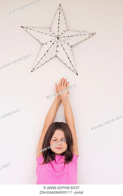Girl reaching toward star shape hanging above her head, eyes closed