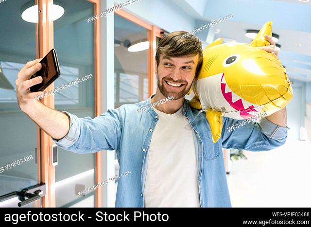 Smiling creative businessman taking selfie with balloon shark in corridor at workplace