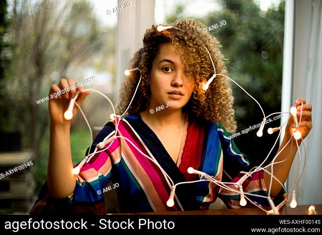 Confused curly haired woman holding illuminated Christmas lights at home