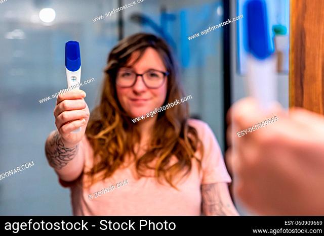 An over the shoulder view as a beautiful caucasian woman stands in front of bathroom mirror with plastic pregnancy test kit showing positive result