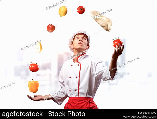 Young male chef juggles with food ingredients. Handsome chef in white hat and red apron in light kitchen interior. Professional cooking classes advertising