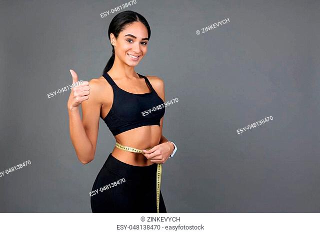 Good results. Positive delighted pleased sportswoman wearing black sport suit keeping her right hand bent in elbow with thumb up while measuring waist