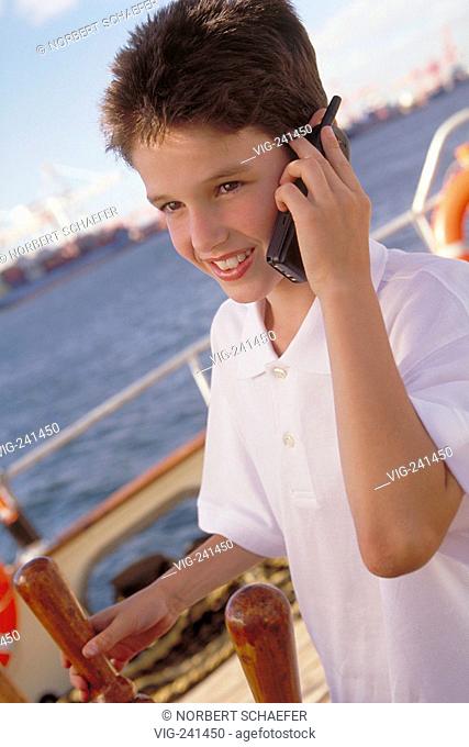 harbour, portrait, 13 years old boy wearing white shirt stands on bord of a boat on sea at the steering wheel making a call with his handy  - GERMANY