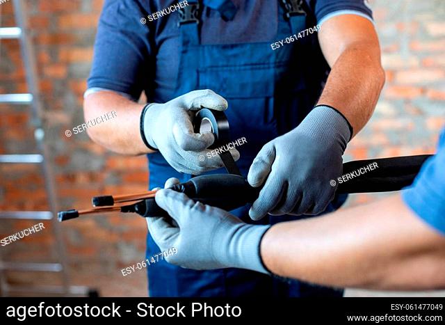 Two adult male workers in overalls are preparing to install underfloor heating pipes. Close up of repairmen hands in work gloves fastening pipes and wires with...