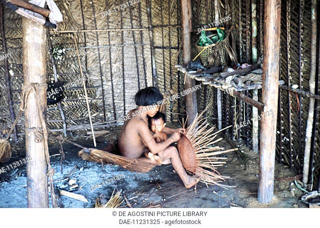 Yanomami woman with a baby on her lap building a straw basket inside a hut, Mato Grosso, Brazil