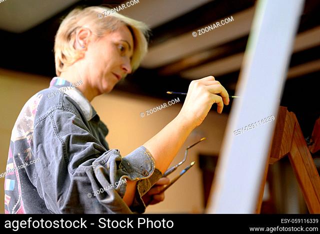 Happy woman paints on canvas using thin brush. Canvas stands on the easel. Artist draws at easel. Side view of female painting picture on canvas in art home...