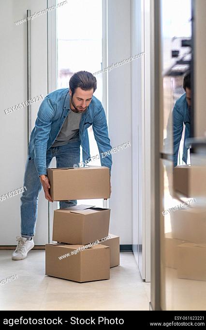Folding boxes. Man standing with his back to door in apartment bending over with box above stack in daylight