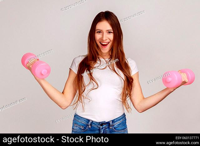 Portrait of excited woman standing with raised arms and holding pink dumbbells, active lifestyle, wearing white casual style T-shirt