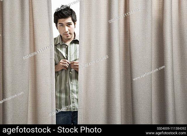 Hispanic man buttoning shirt behind curtain