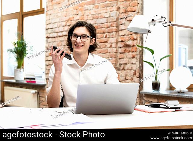 Happy businessman talking on smart phone through speaker sitting at desk in office