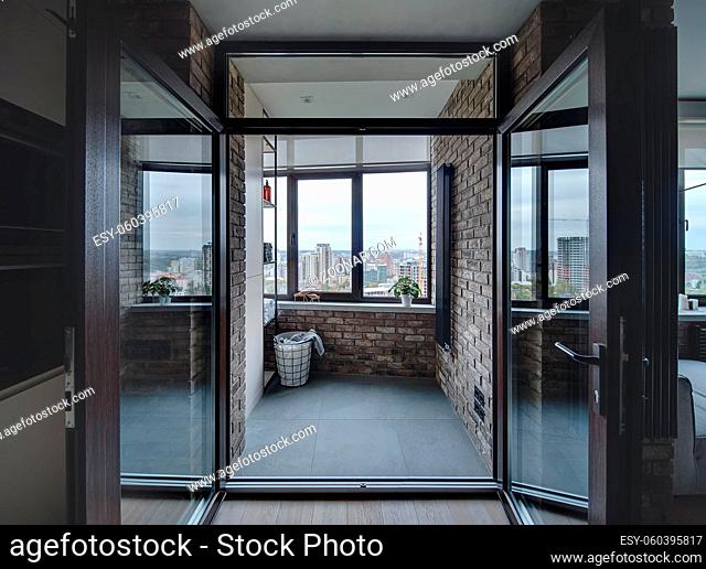Entrance to the loft style balcony with brick walls and dark tiles on the floor. There is shelves with towels and bottles, basket with clothes, glass doors