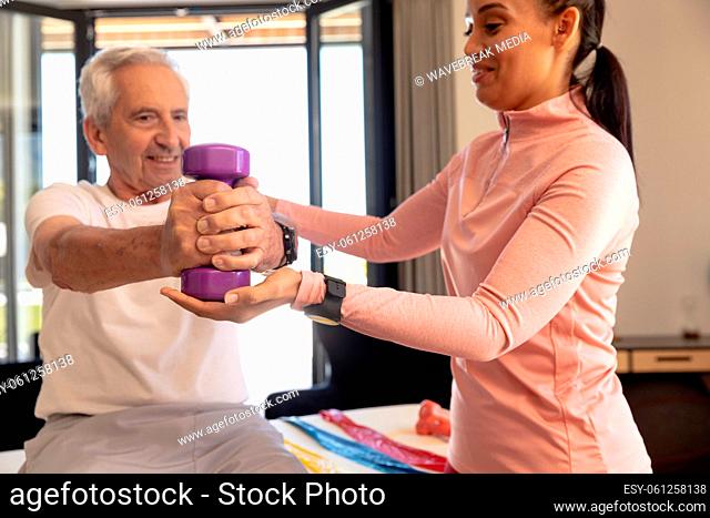 Smiling biracial female physiotherapist helping caucasian senior man in lifting dumbbell at home